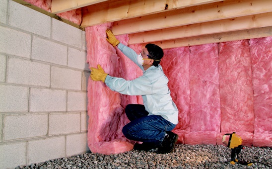 Technician installing pink fiberglass batt insulation in a crawl space.